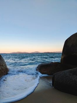 Serene beach scene in SC, Brazil during sunset with waves crashing on rocks and a clear sky.