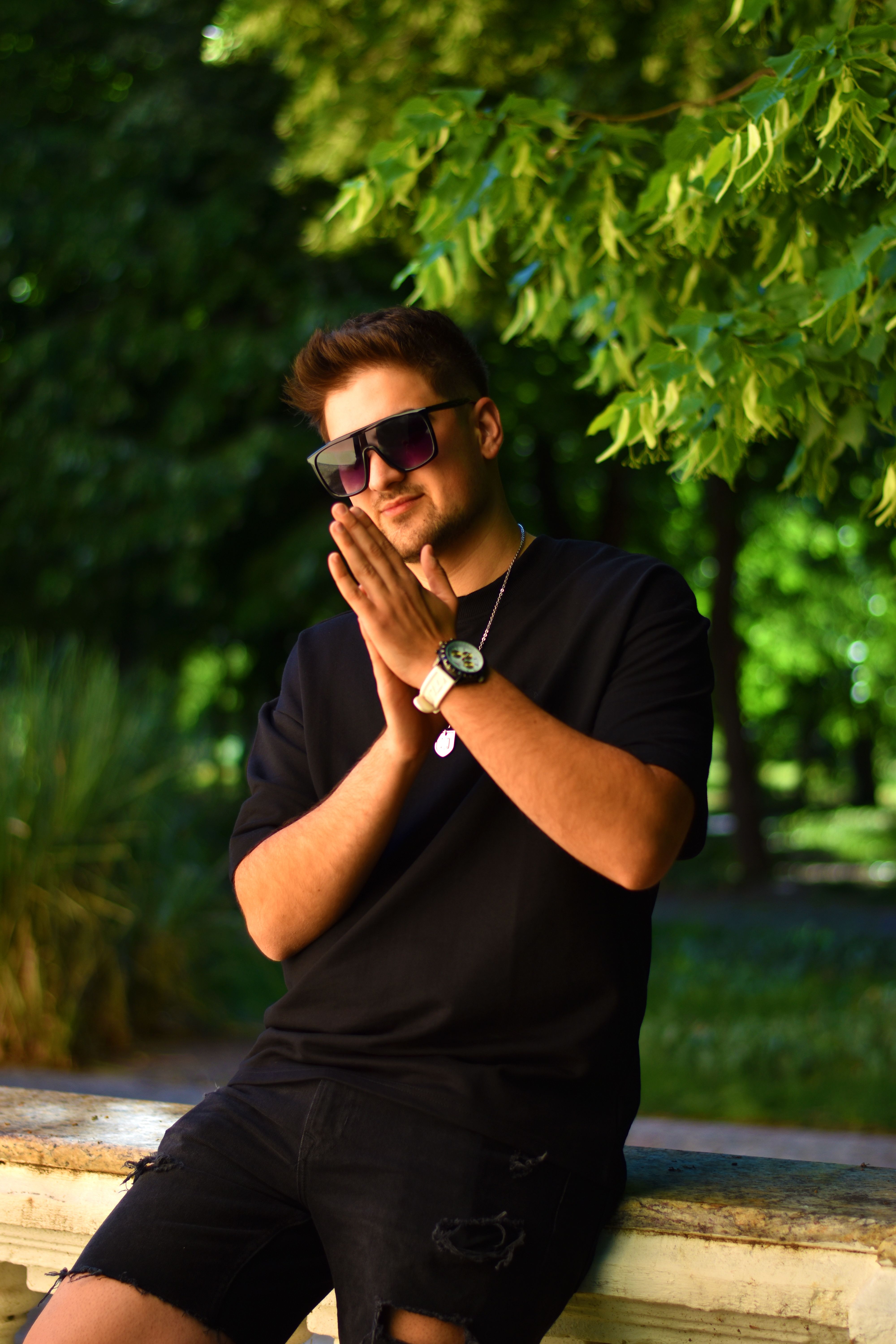 Man wearing sunglasses and a black t-shirt, posing outdoors in a park setting.