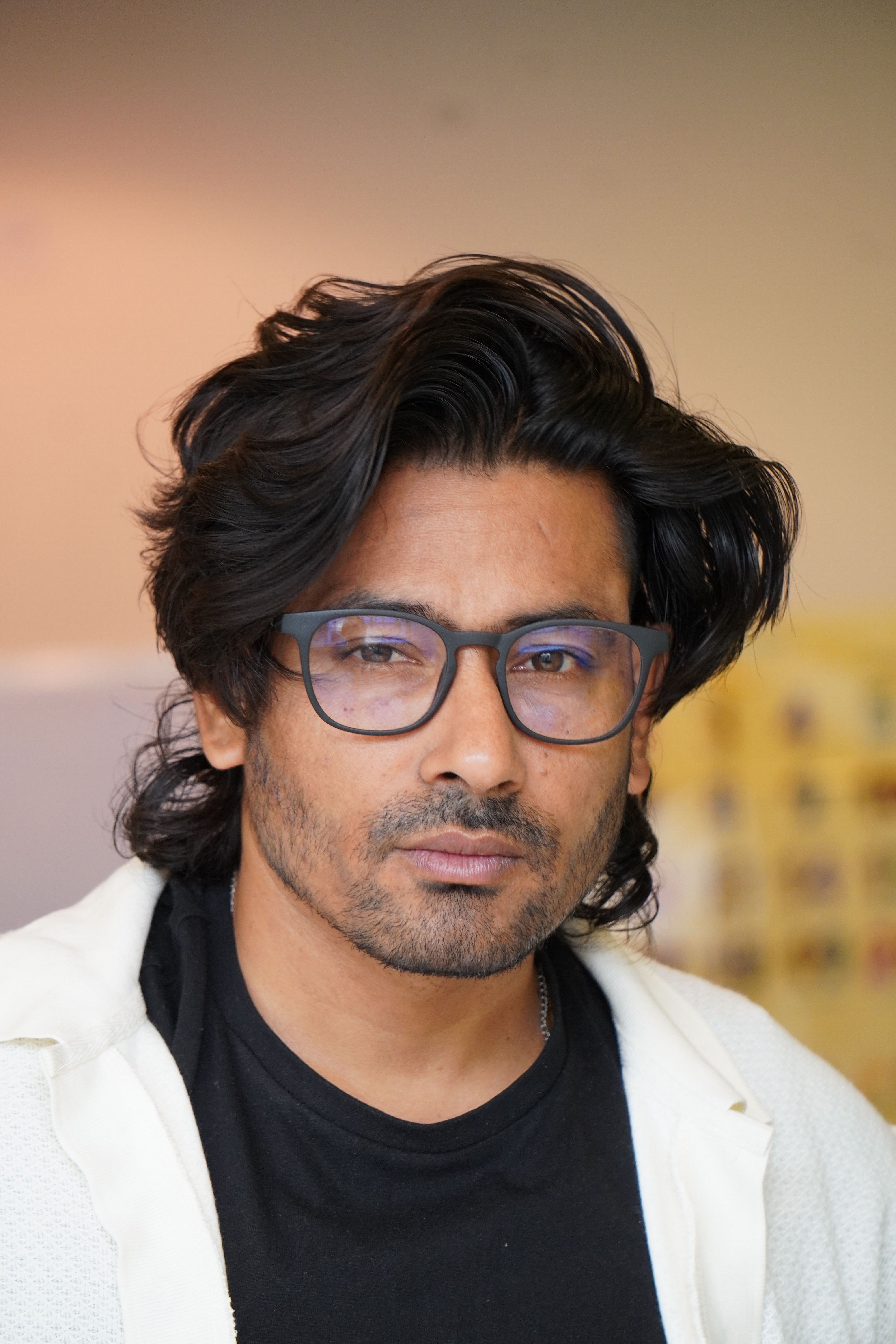 Portrait of a man with black hair and eyeglasses, looking contemplative indoors.