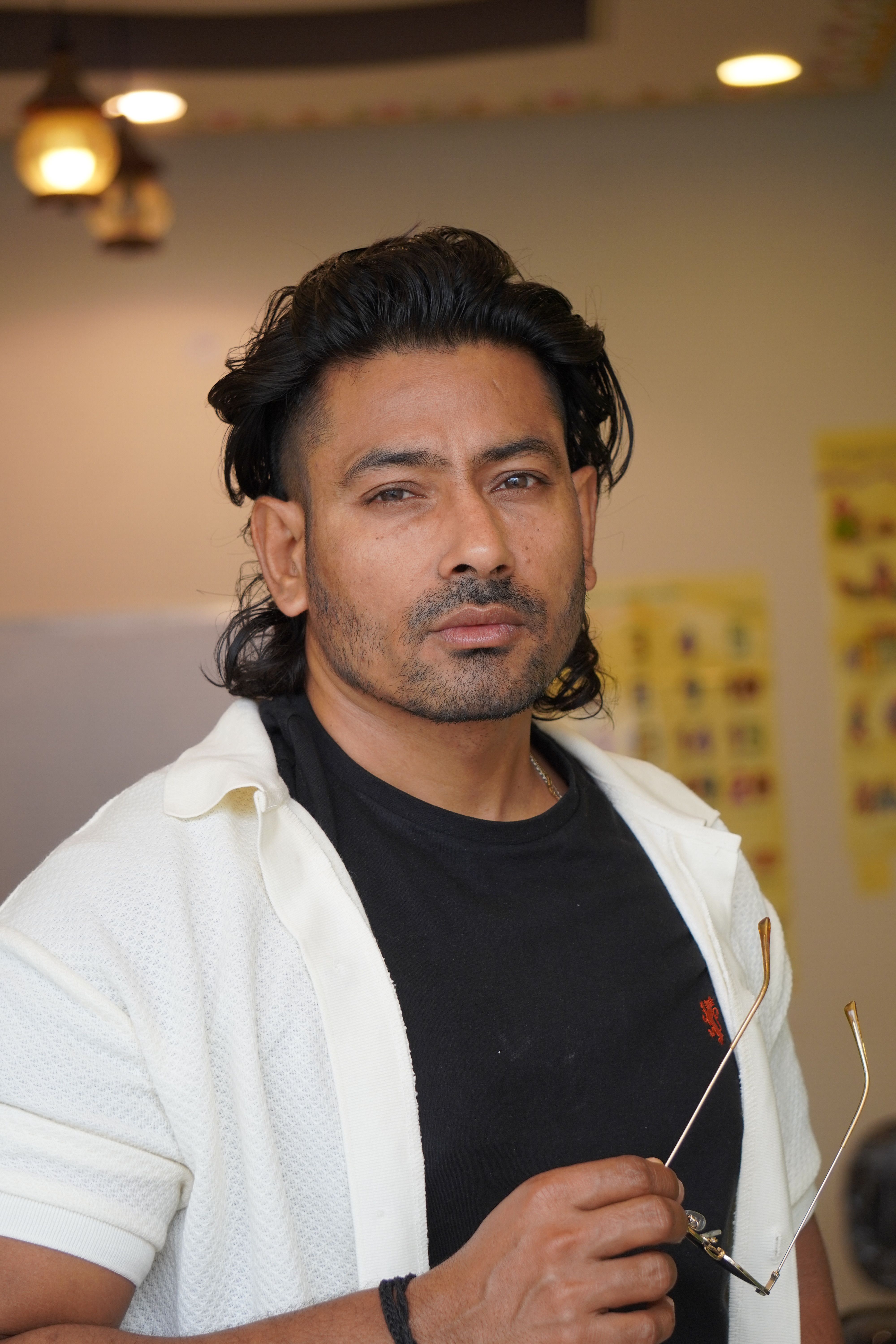 Stylish man in a white shirt and black tee stands indoors, holding glasses with a confident expression.