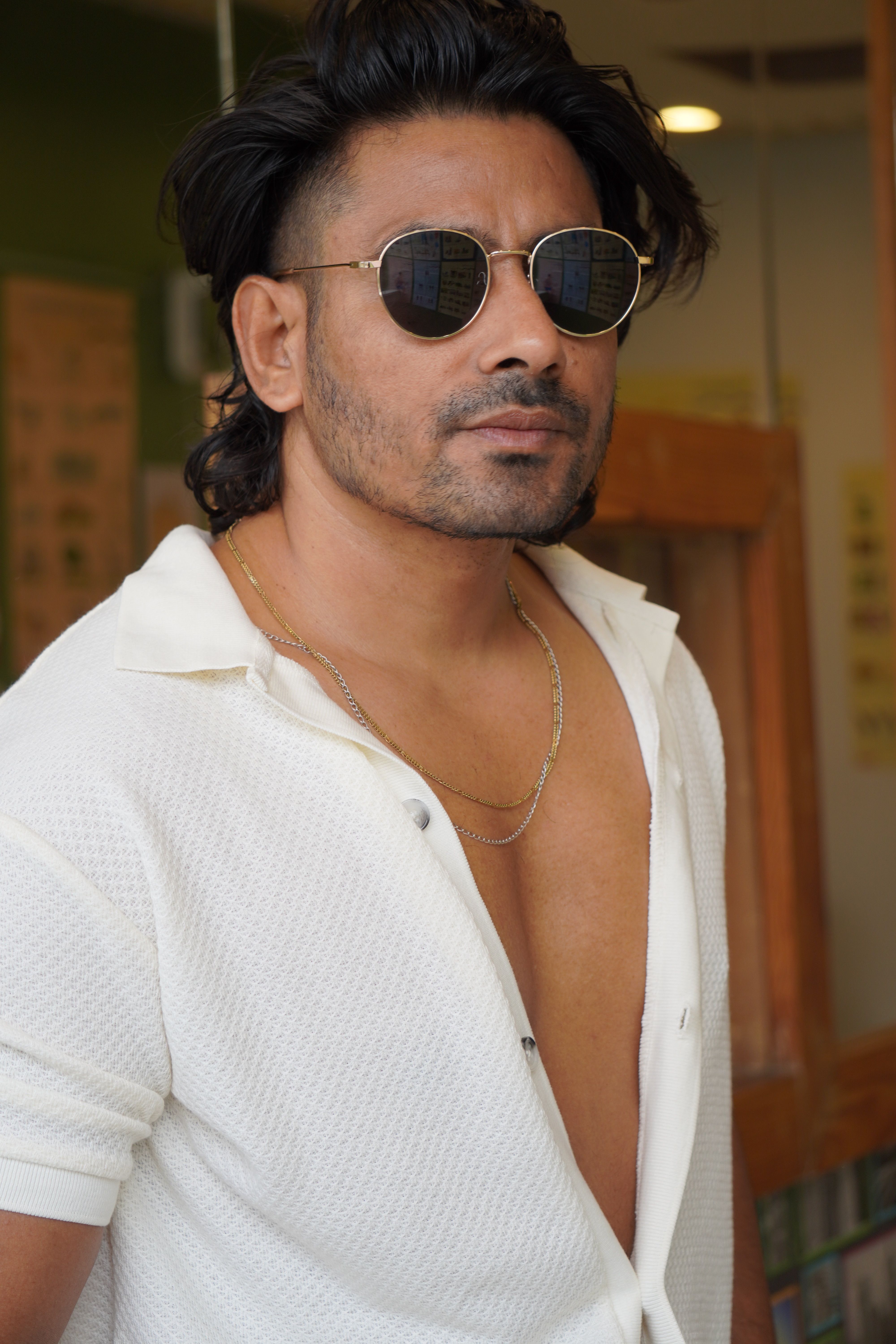 Portrait of a confident man indoors wearing sunglasses and a white shirt with an open collar.