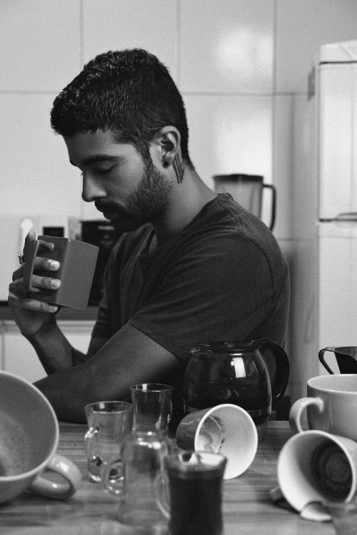 Free Thoughtful man sitting in a kitchen, holding a coffee mug, surrounded by dishes in a black and white photograph. Stock Photo