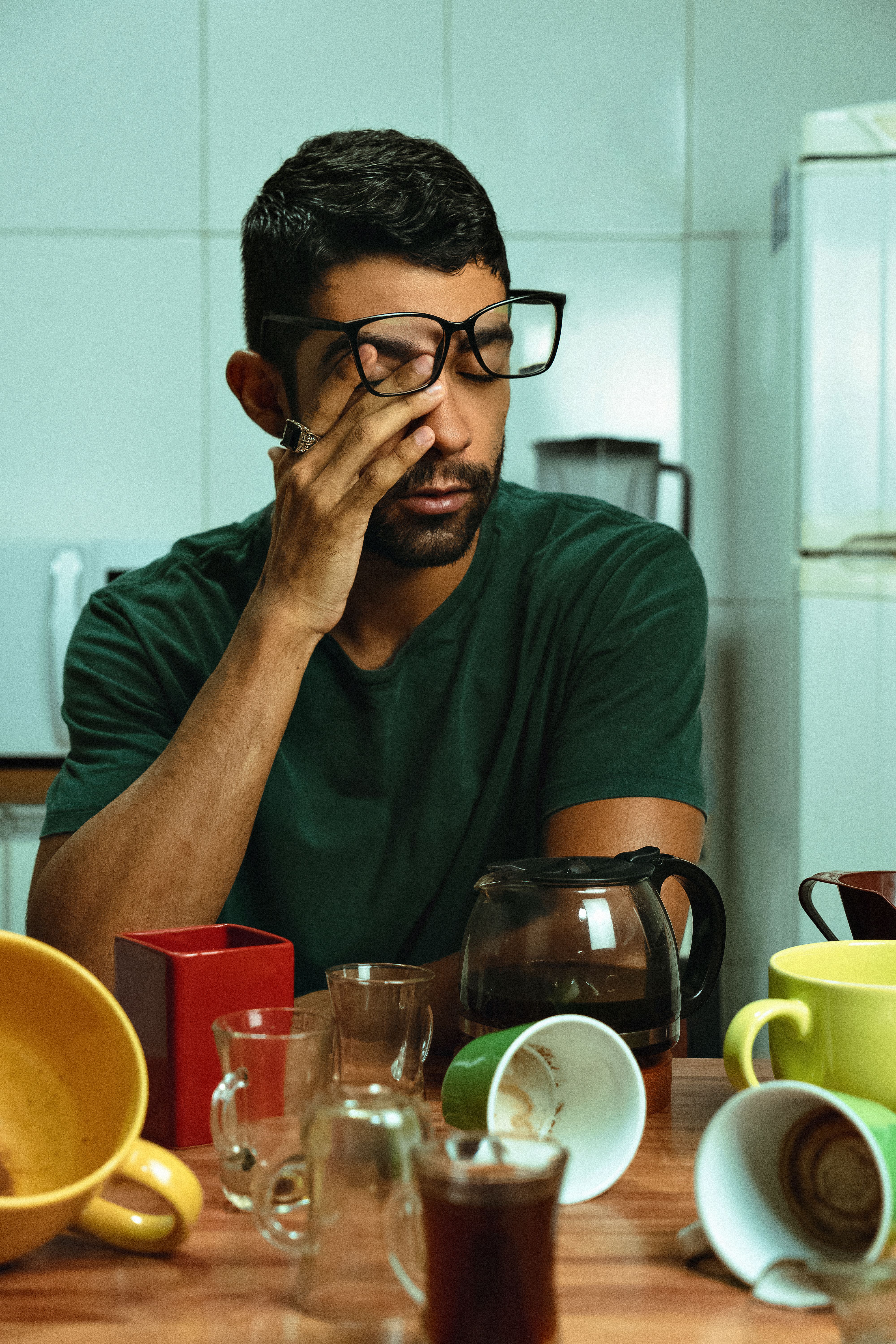 Man feeling tired, surrounded by empty coffee cups in a kitchen. Concept of exhaustion and caffeine dependence.