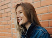 一位长发女子，身穿牛仔夹克，倚靠在砖墙上，面带微笑