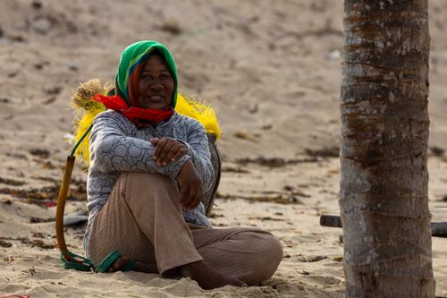 Free A cheerful woman with a colorful hood sits on a sandy beach, smiling in the sunlight. Stock Photo