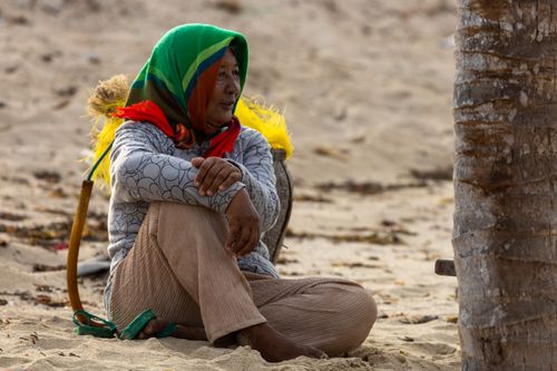 Free A senior woman in a colorful headscarf sits on the sandy beach beside a palm tree, enjoying a peaceful moment. Stock Photo