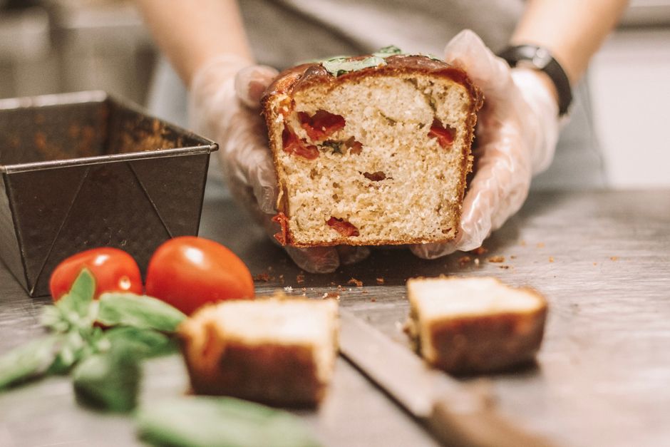 A homemade loaf of tomato herb bread being sliced on a kitchen counter surrounded by fresh tomatoes and basil.