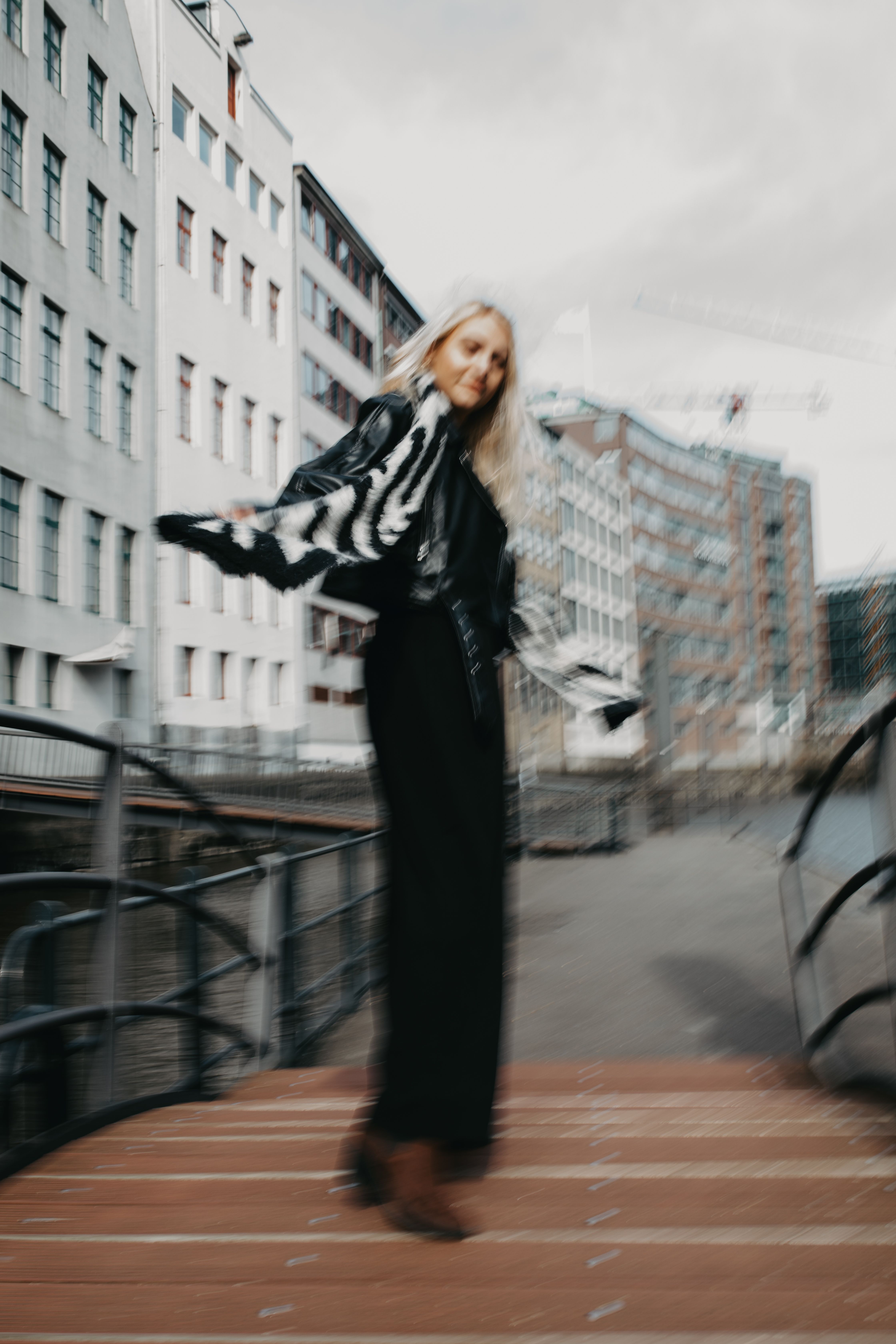 A stylish blonde woman in motion on a wooden bridge in a city, featuring urban architecture in the background.