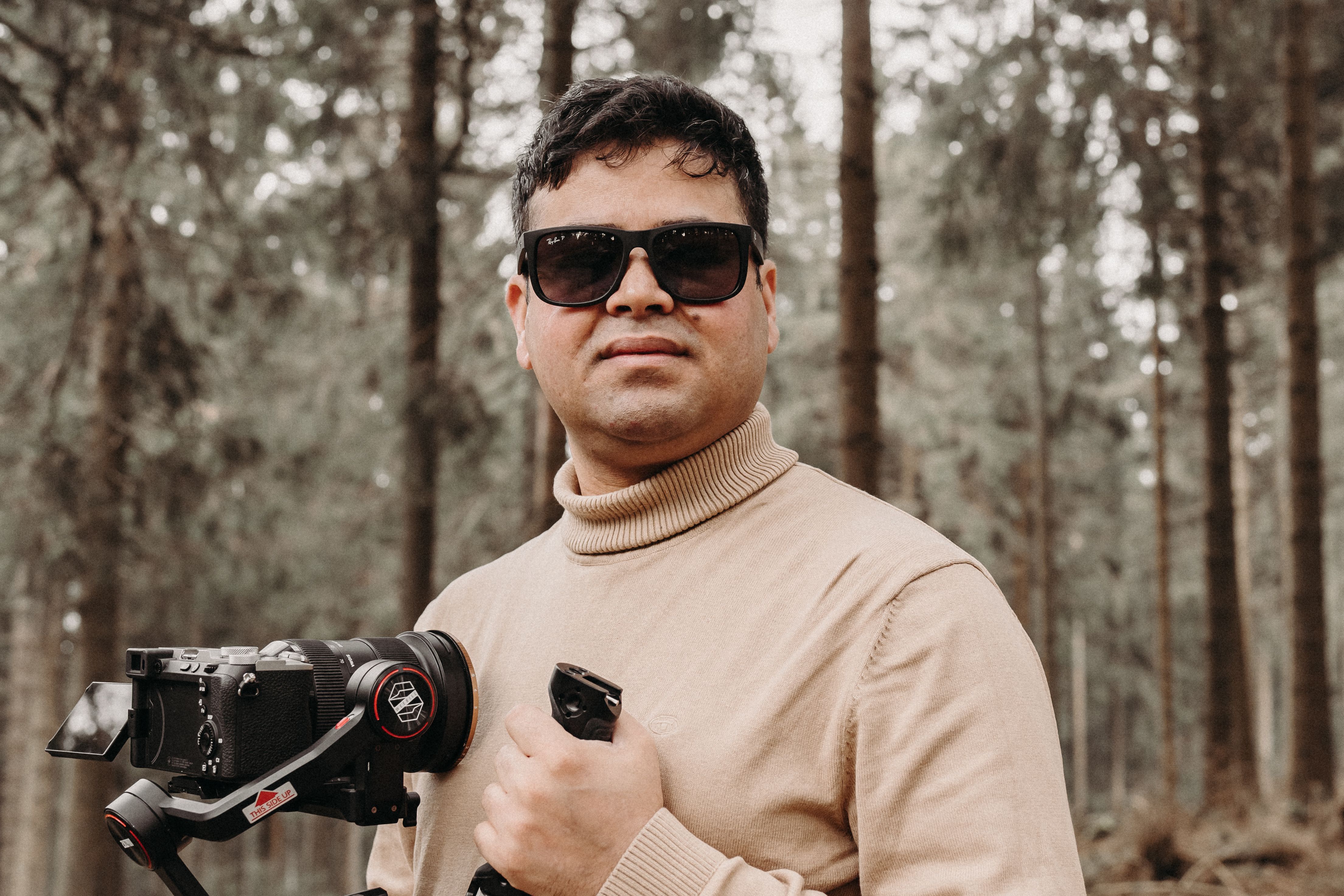 Man with camera and gimbal in a forest, wearing sunglasses and a turtleneck.