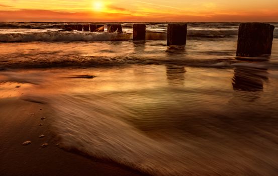 Beautiful sunset with waves crashing on a serene beach and wooden pier.