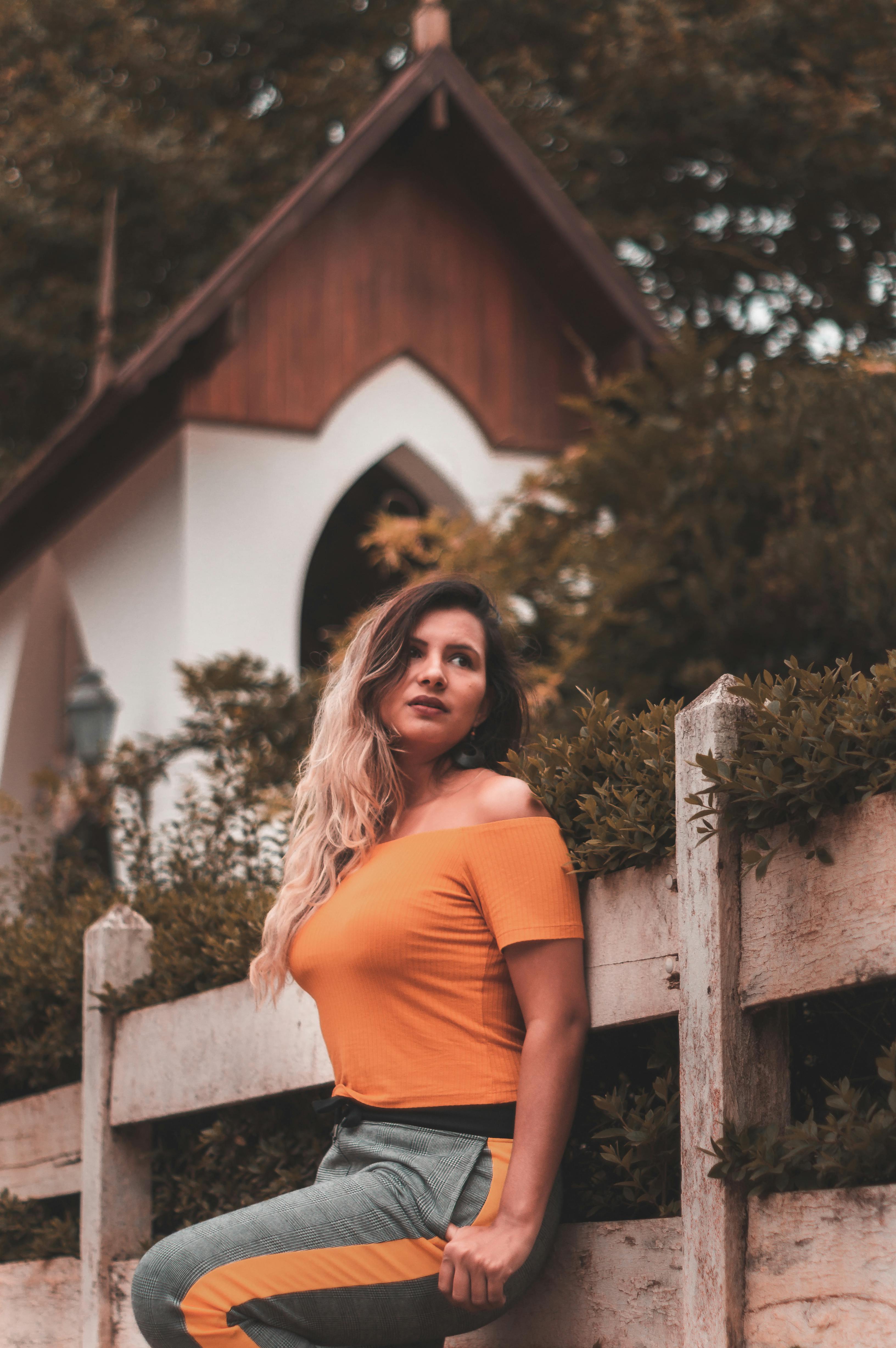 一位身穿橙色上衣的时尚女子在木栅栏旁摆姿势，背景是一座乡村建筑