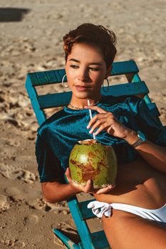 Stylish woman enjoying a fresh coconut drink on a sunbed at the beach.