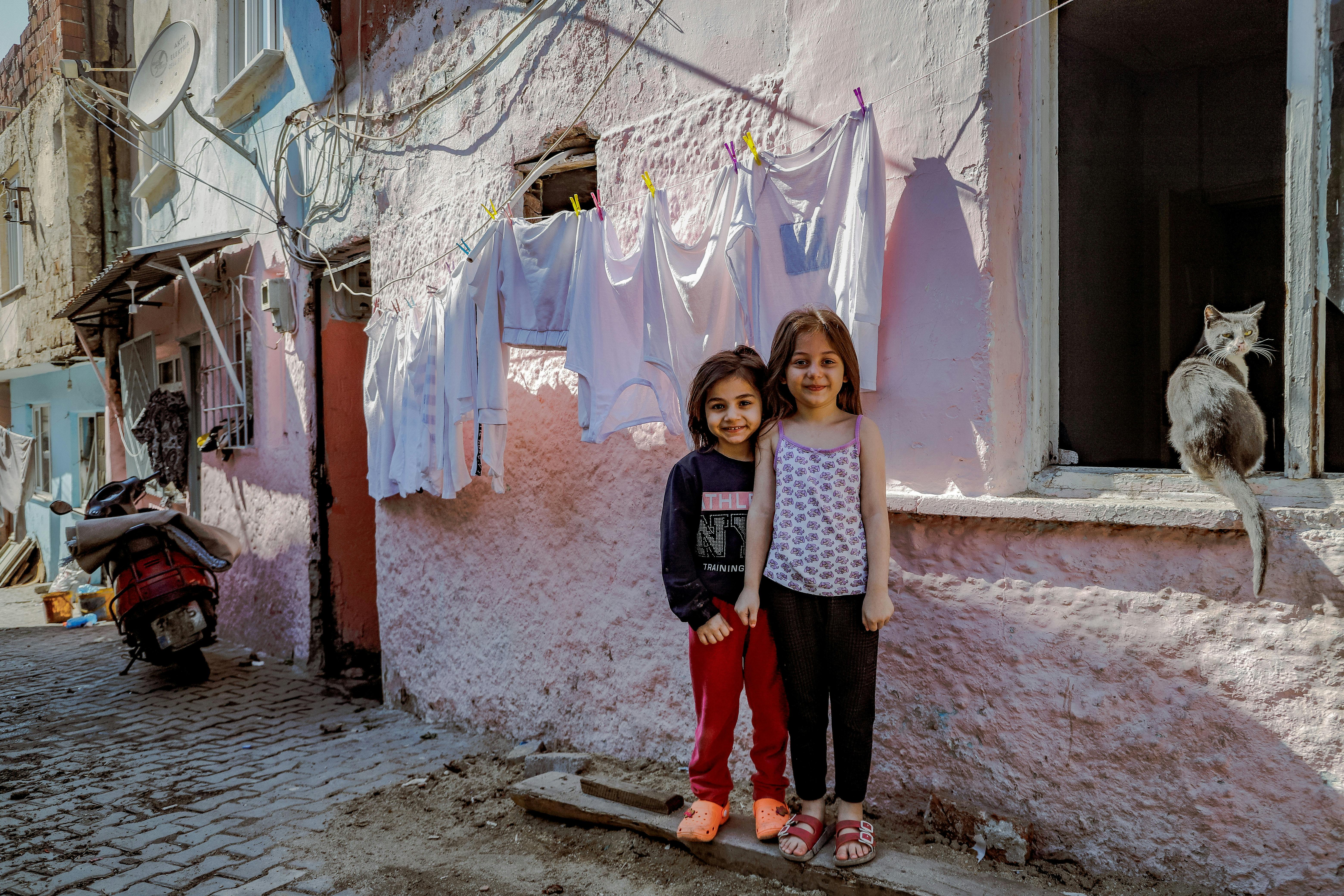 Two smiling girls stand outside in an Istanbul street, with a cat and hanging laundry visible.