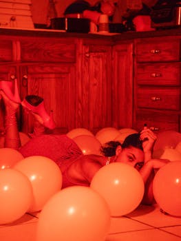 Moody indoor portrait of a young woman in red lighting, surrounded by balloons on a kitchen floor.