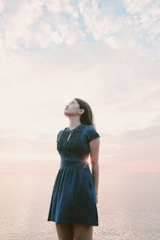 一名身穿蓝色连衣裙的女子若有所思地抬起头，站在海边看着日落