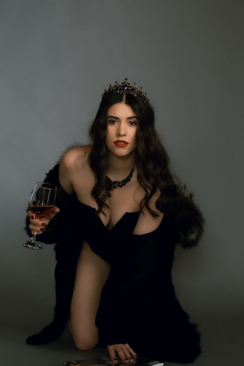 Free A woman with a crown in a black coat kneels elegantly with a glass of wine in a studio. Stock Photo
