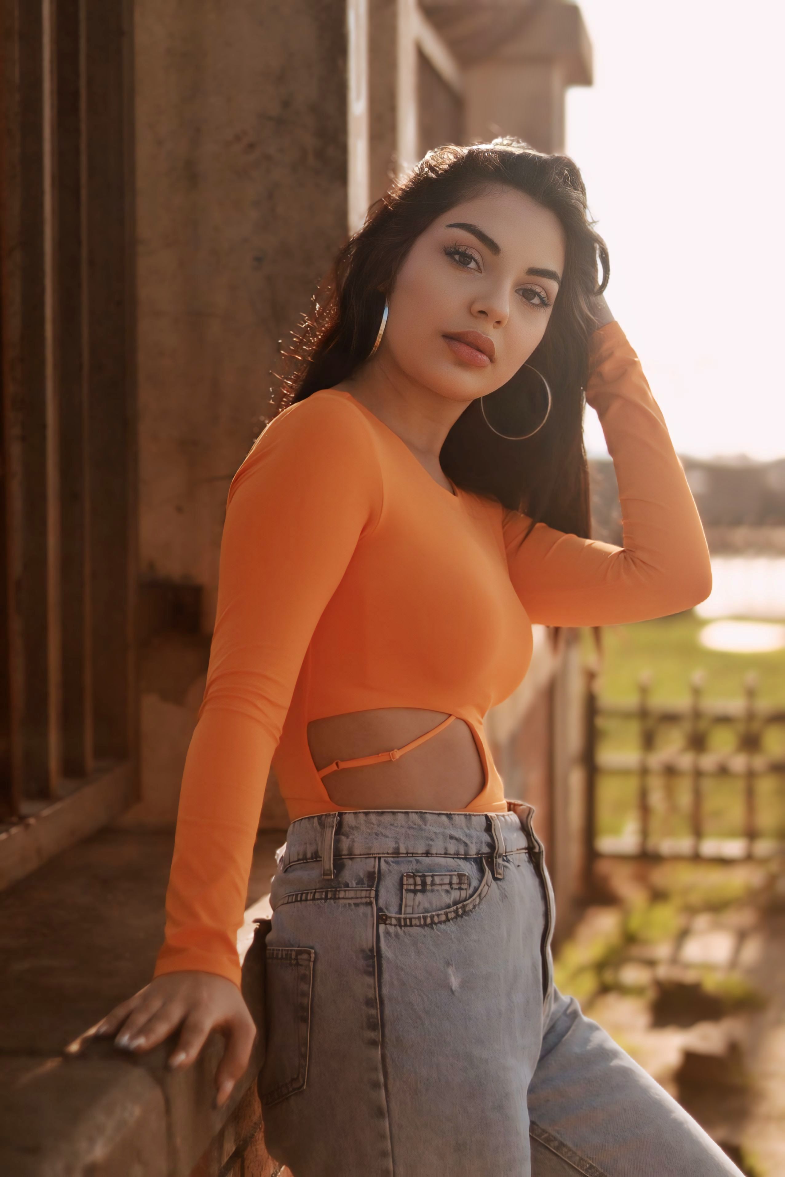 Stylish young woman with long hair poses confidently in bright orange blouse outdoors.