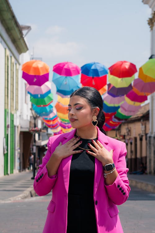 A woman in a pink blazer and black dress standing in the middle of a street with colorful umbrellas