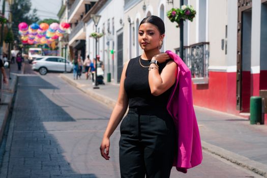 Stylish woman in a black outfit and pink jacket strolling an urban street adorned with colorful decorations.