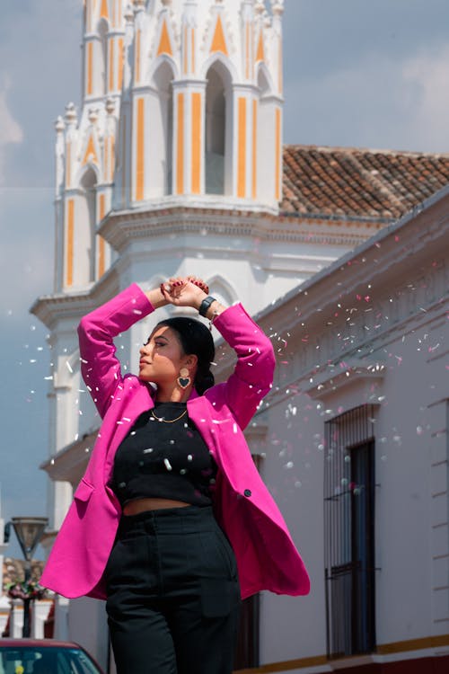 Confetti around Woman in Pink Suit Jacket Standing with Arms Raised and with Church behind