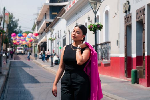 Elegant woman in a pink jacket walking down a vibrant street with colorful decorations.