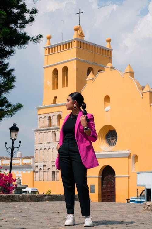 A woman in a pink blazer and black pants stands in front of a yellow church