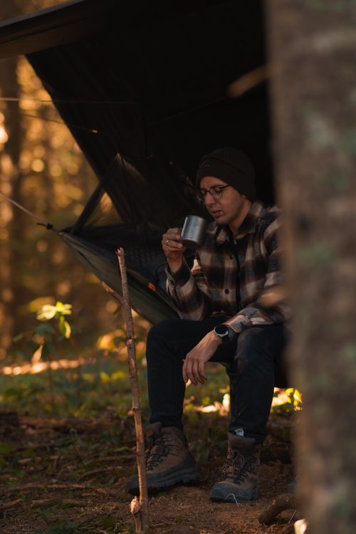 Free A man wearing a beanie and plaid shirt sipping coffee in a cozy forest camp setup. Stock Photo
