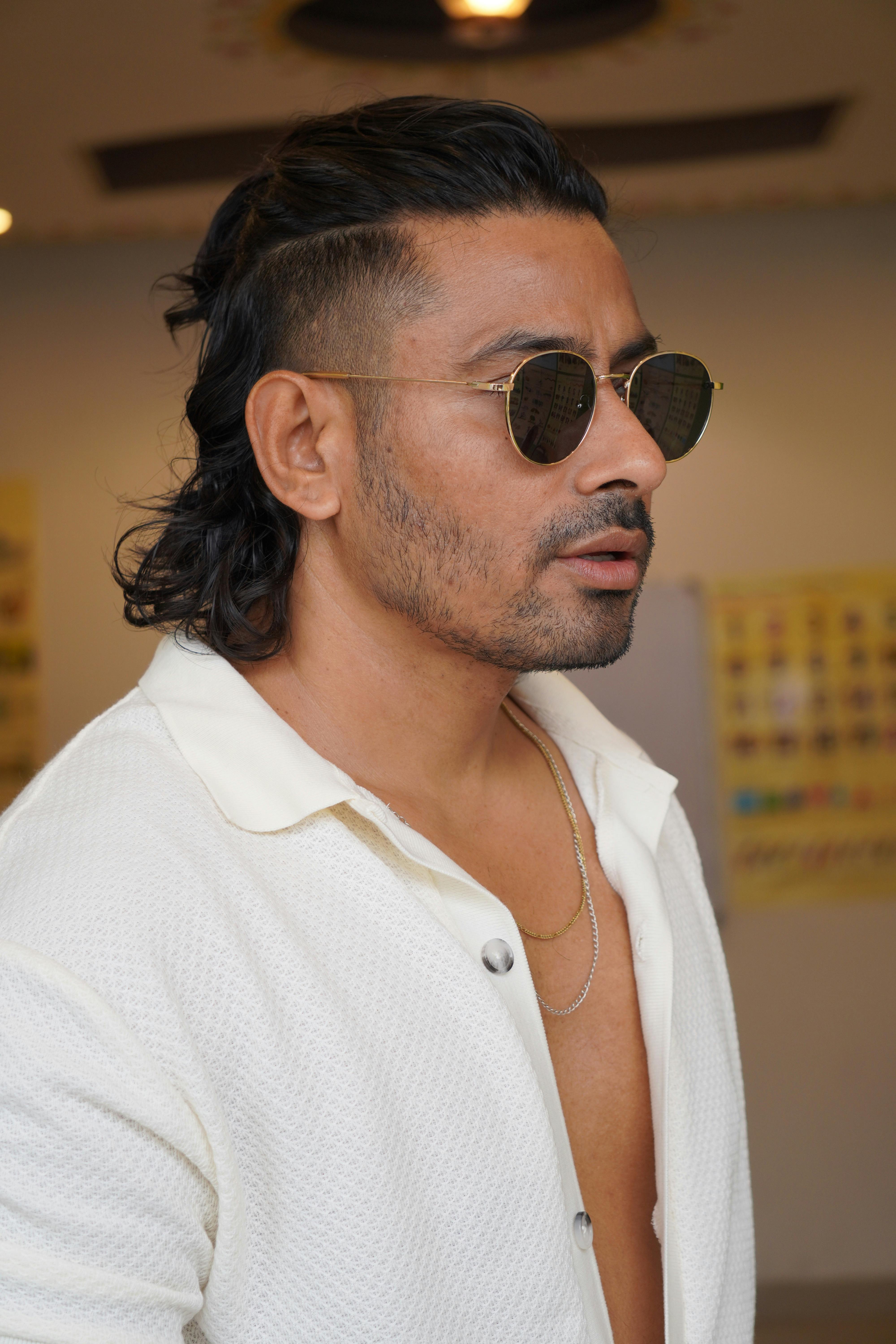 Fashionable man with sunglasses and long hair posing indoors for a portrait.
