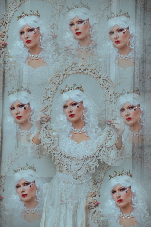 Free A regal woman in a crown and white gown reflected multiple times in an ornate mirror, conveying elegance and fantasy. Stock Photo