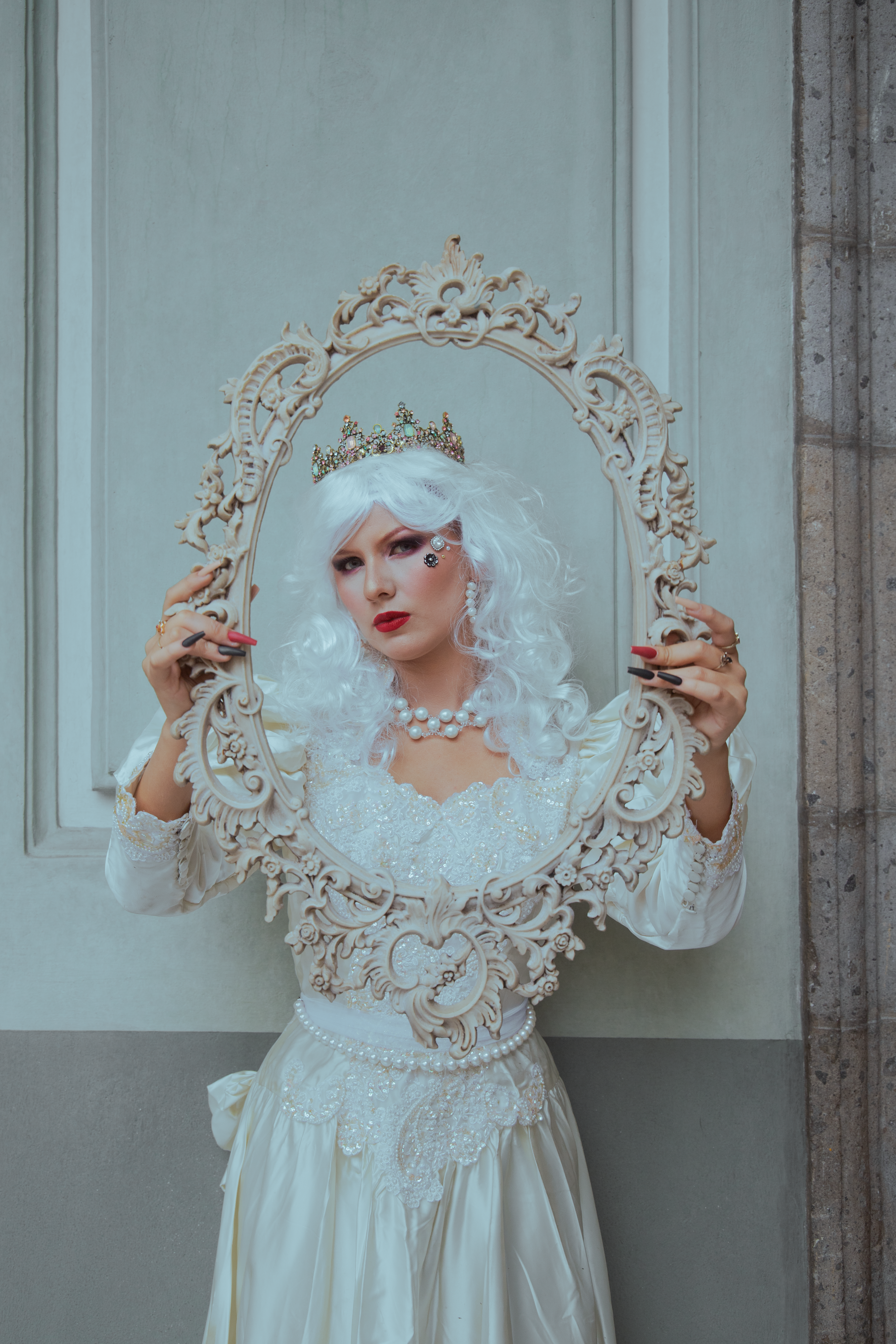A woman in vintage attire and a crown holds a decorative frame, exuding elegance and classic charm in an indoor setting.
