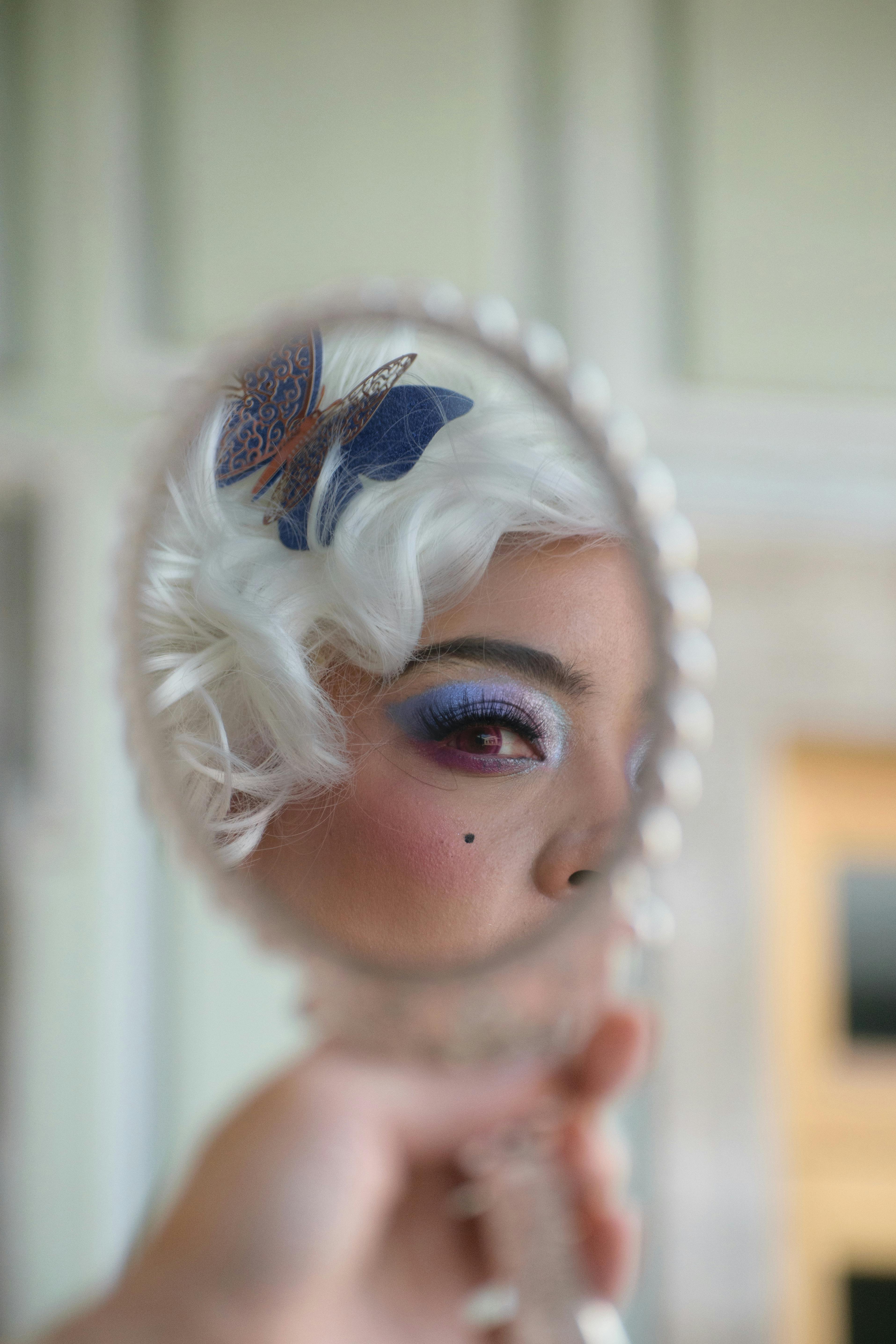 A stylish close-up of a woman's reflection in a handheld mirror with white hair and colorful makeup.