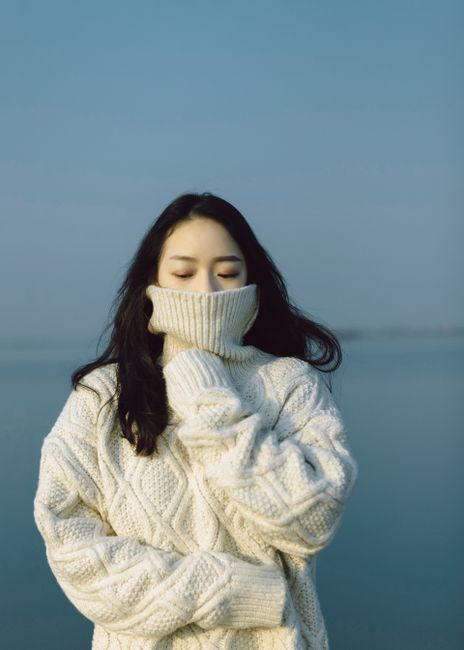 Woman wearing a cream knit sweater standing on a breezy beach with a serene sea backdrop.