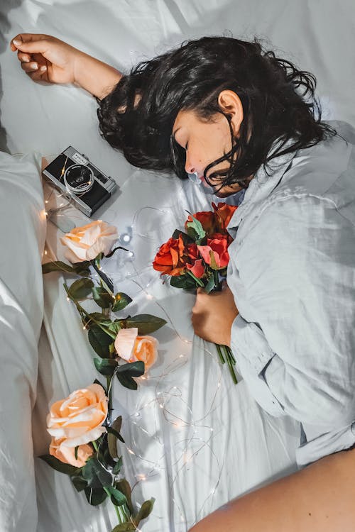 Free High angle side view relaxed sensual female in shirt with bouquet of red aromatic roses lying on comfy cozy bed with photo camera and garland Stock Photo