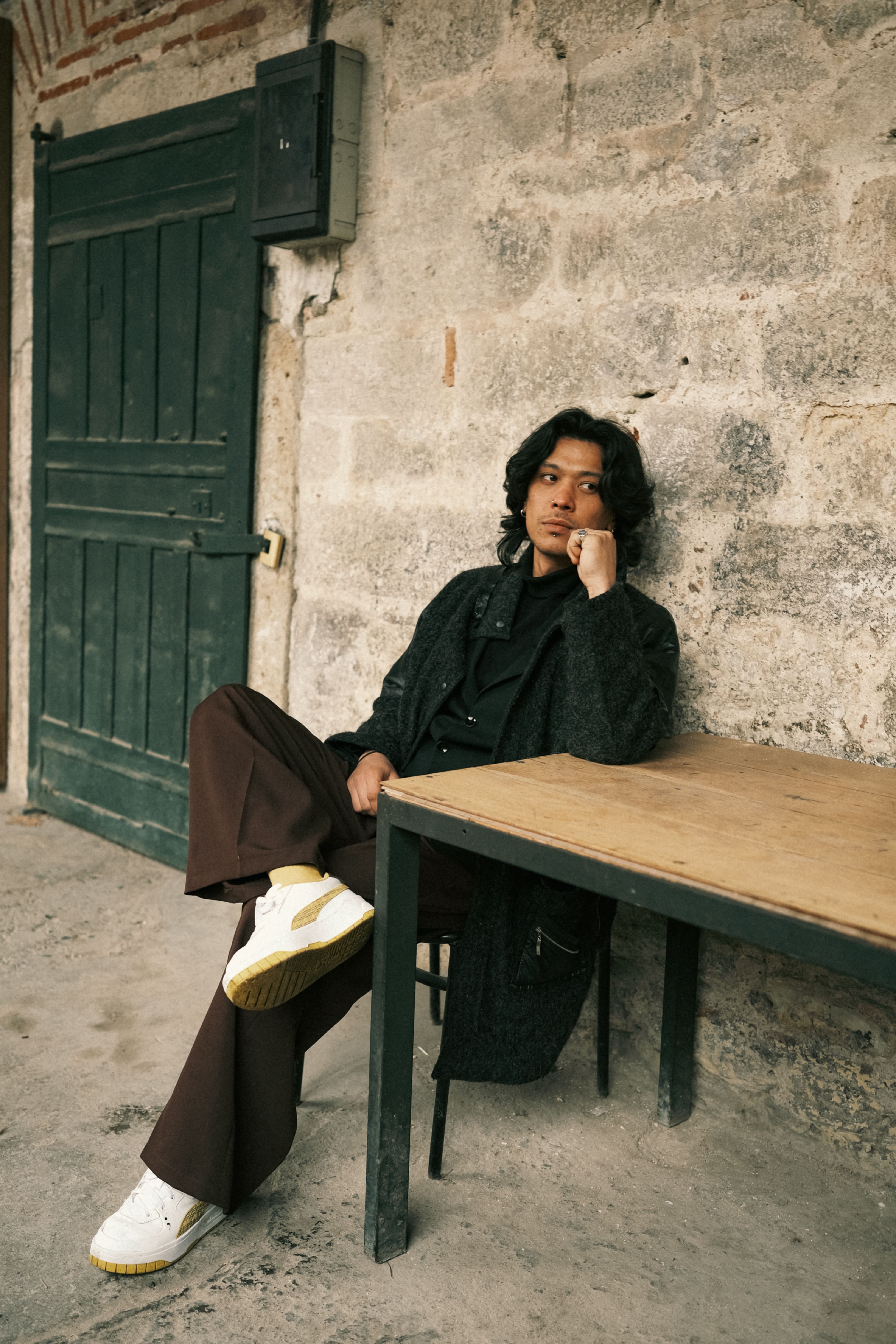 Young man in black clothing seated near a rustic stone wall, exuding a contemplative vibe.