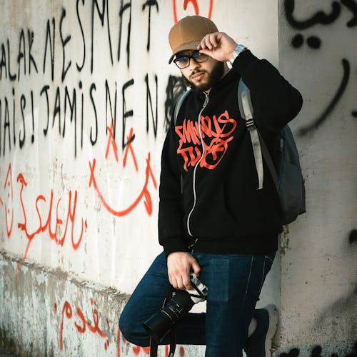 A man with a backpack and a skateboard leaning against a wall with graffiti