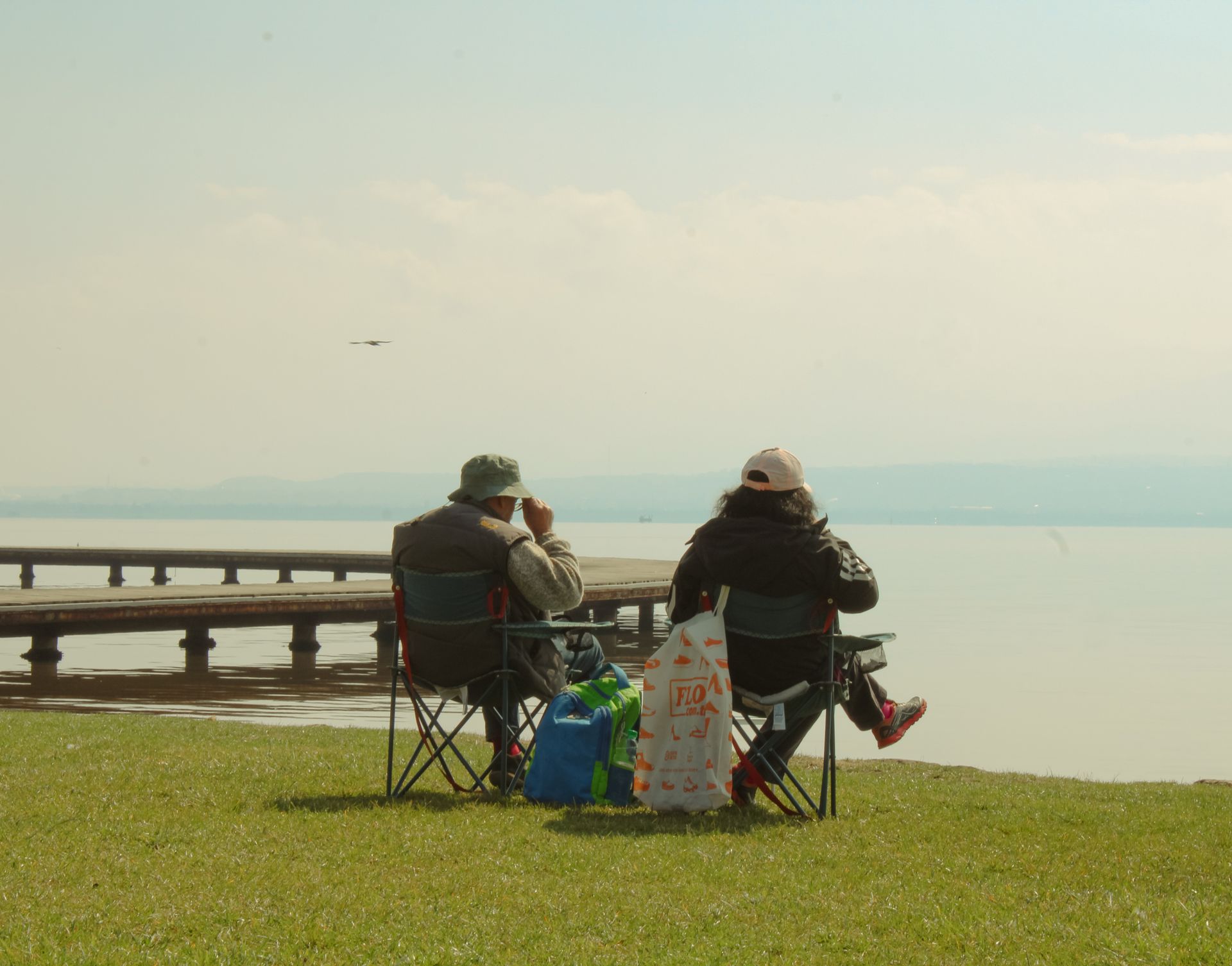 A couple enjoys leisure time sitting on folding chairs by a serene lake with a wooden pier.