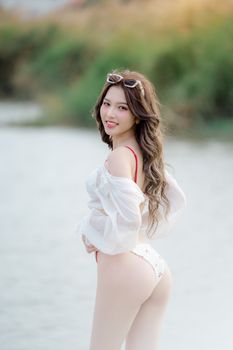Smiling Asian woman in a bikini with sunglasses at a sunny beach, radiating joy.