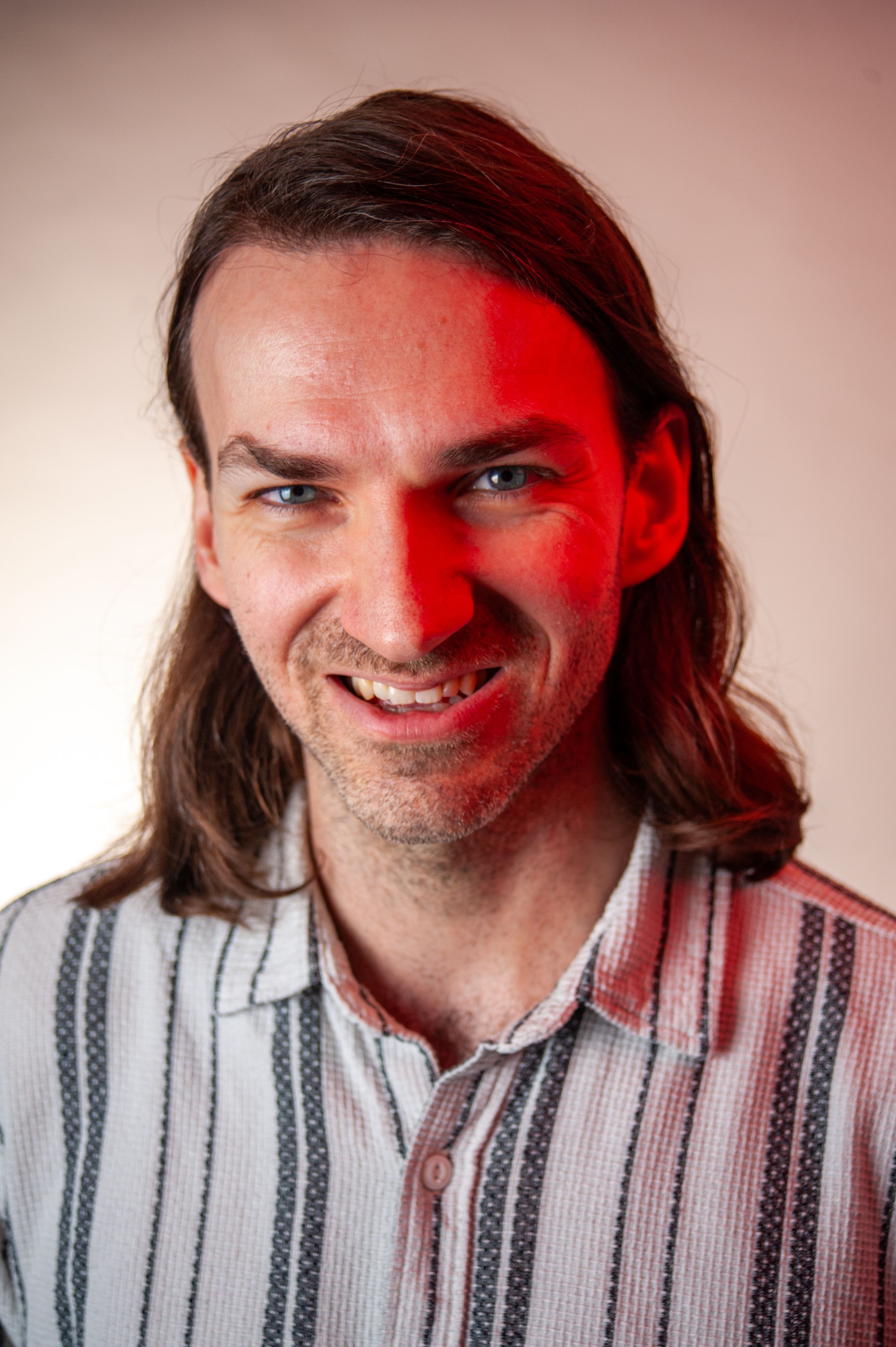 A smiling man with long hair and a patterned shirt, highlighted by red lighting, poses for a portrait.