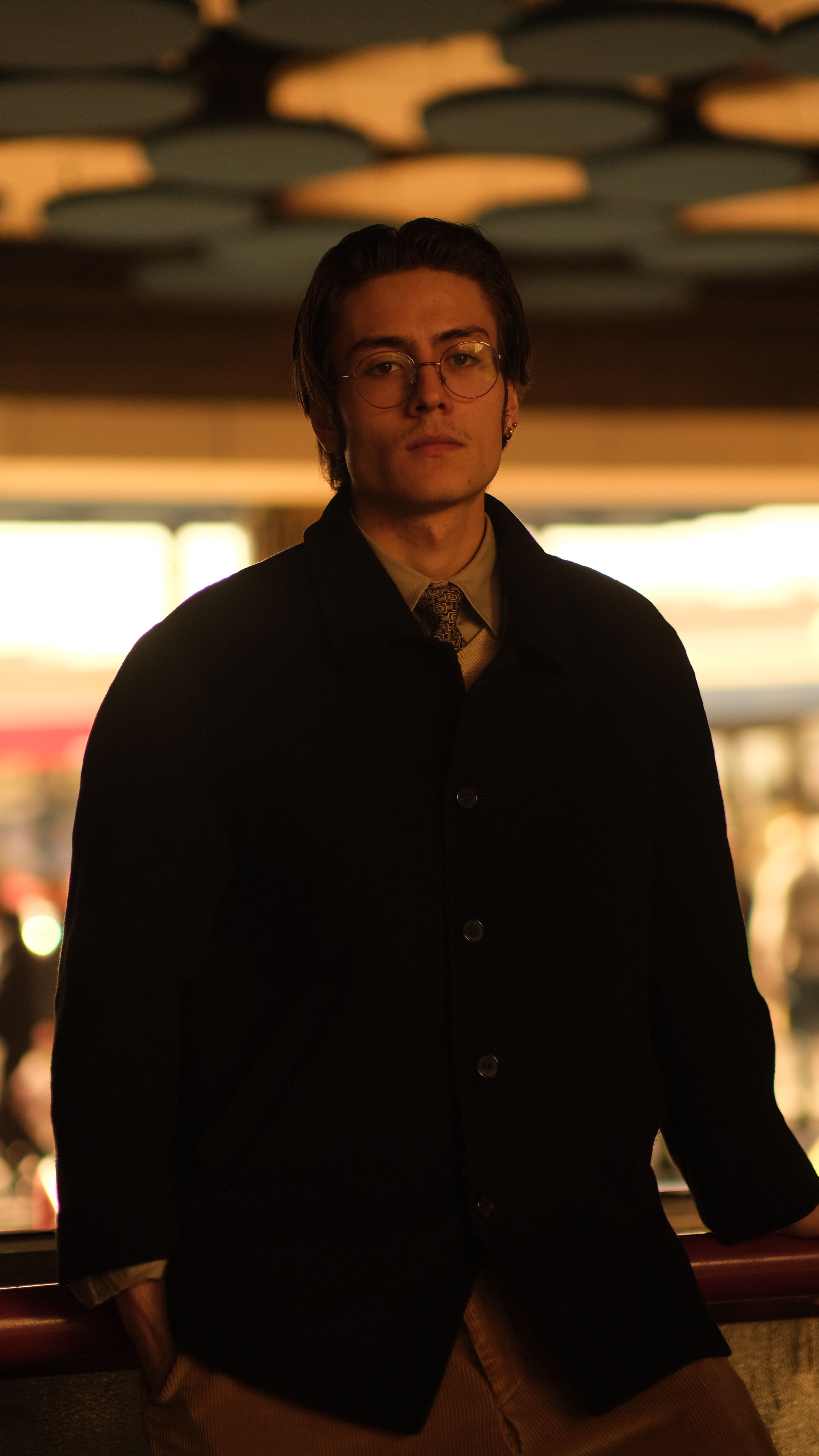 A stylish man poses indoors wearing a jacket, glasses, and necktie, exuding confidence.