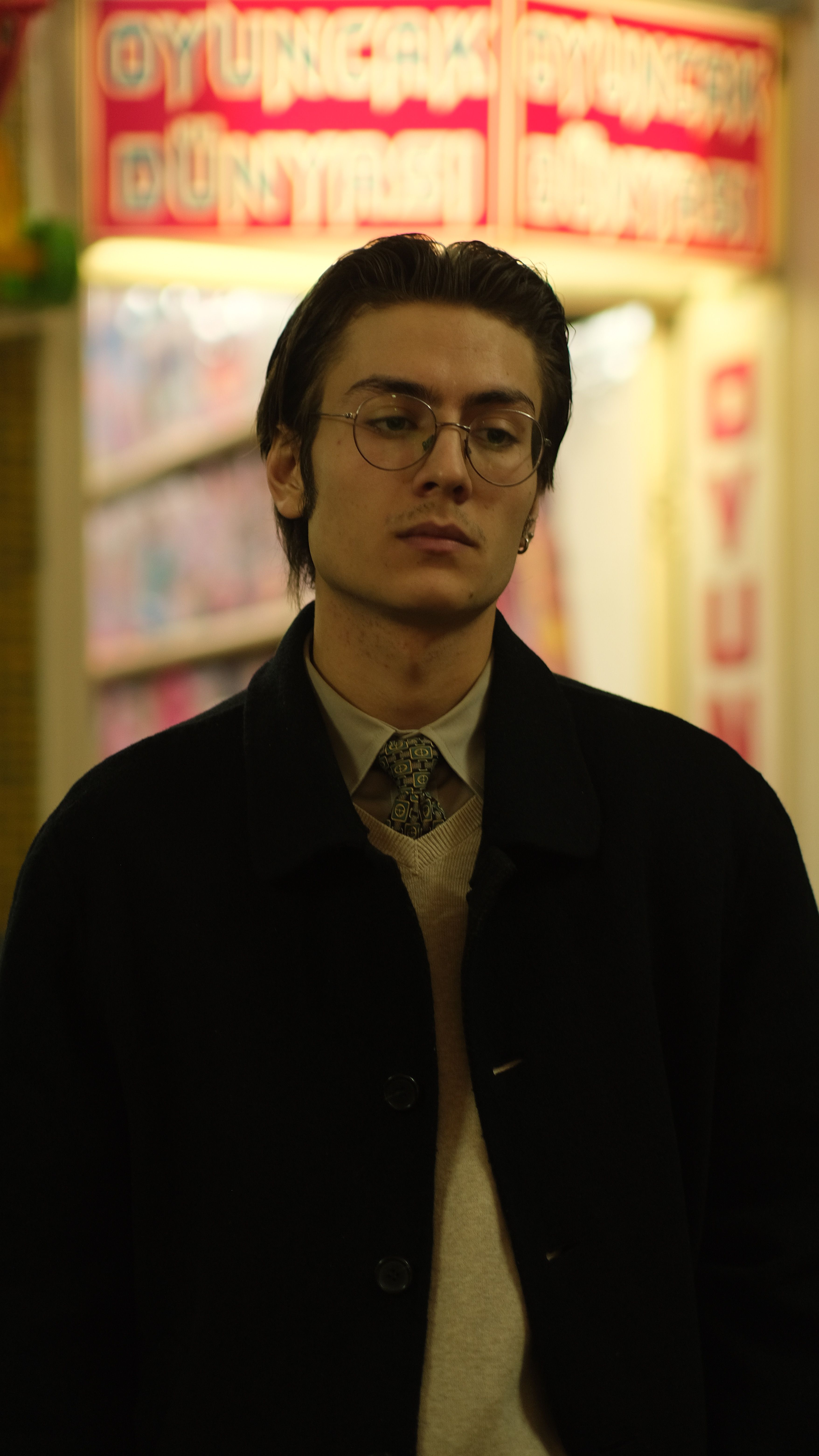 A young man wearing glasses and a jacket poses by neon lights at night.