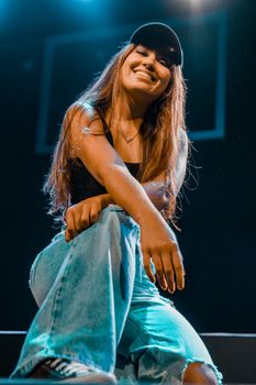 Young woman in casual attire sitting and smiling under stage lights.