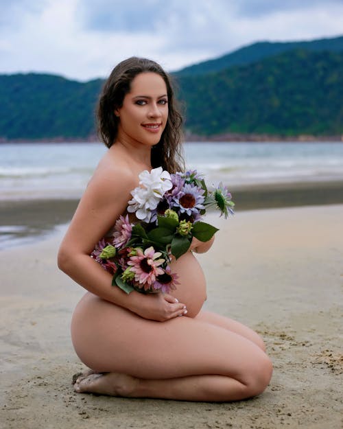A pregnant woman is sitting on the beach holding flowers