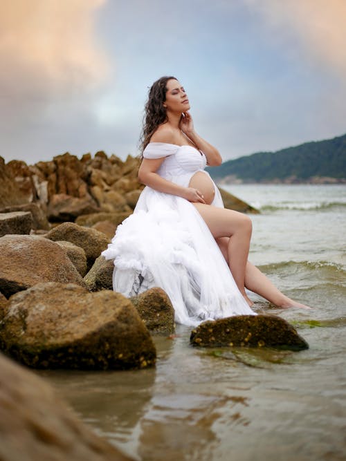 A pregnant woman in a white dress sitting on rocks