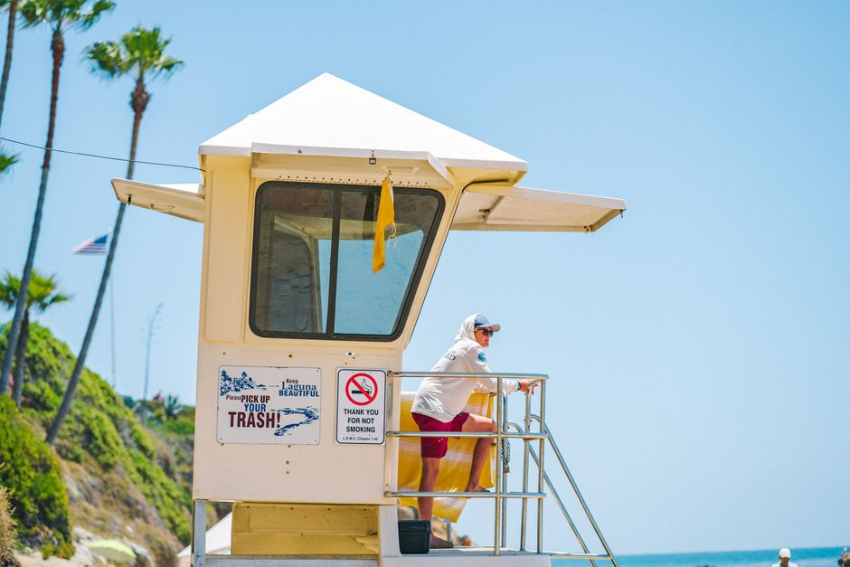 A lifeguard wearing a cap and hoodie watches over a sunny beach from a tower.