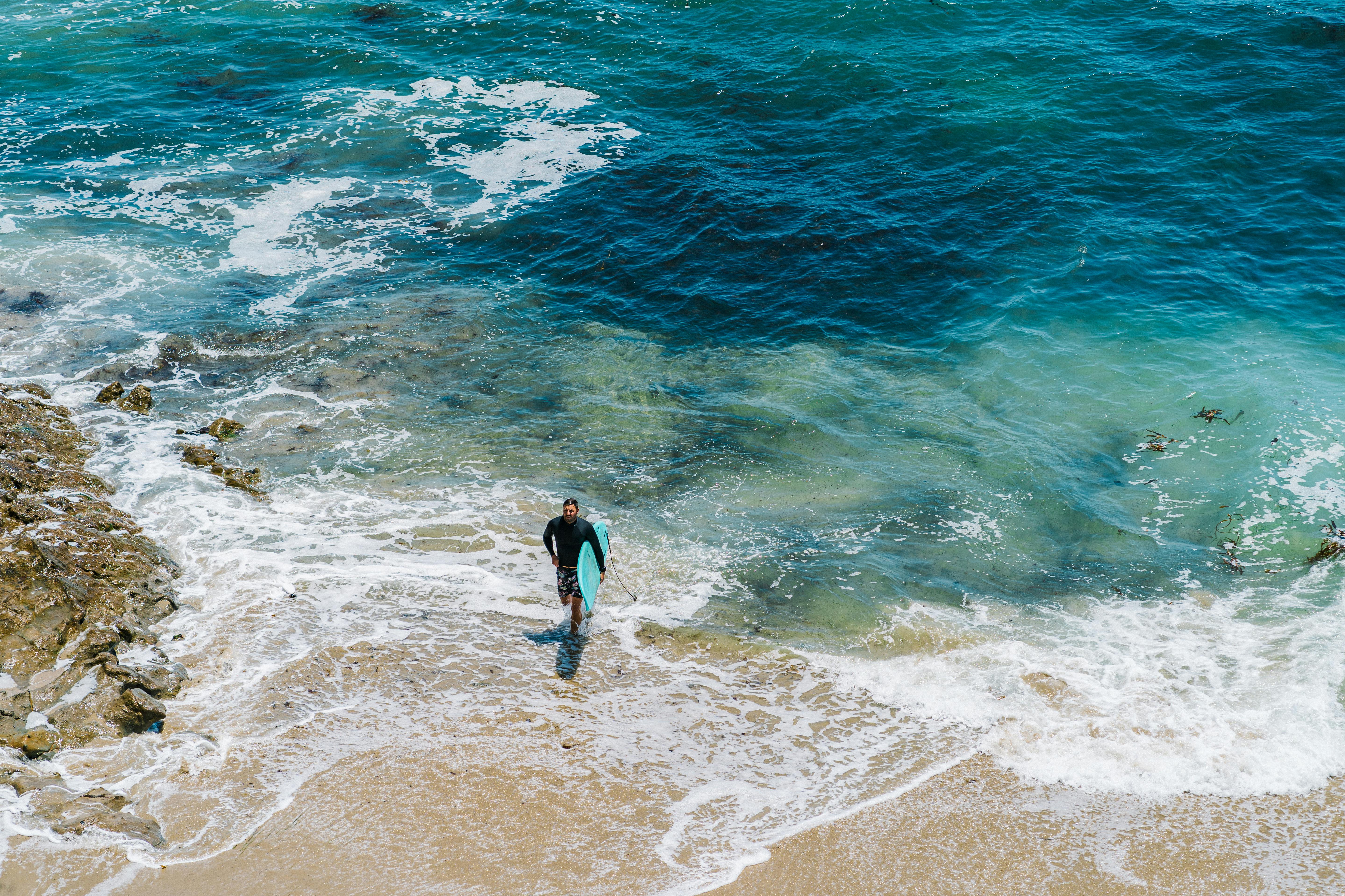 A young man carrying a surfboard walks along a rocky, colorful shoreline, showcasing the vibrant ocean waves.
