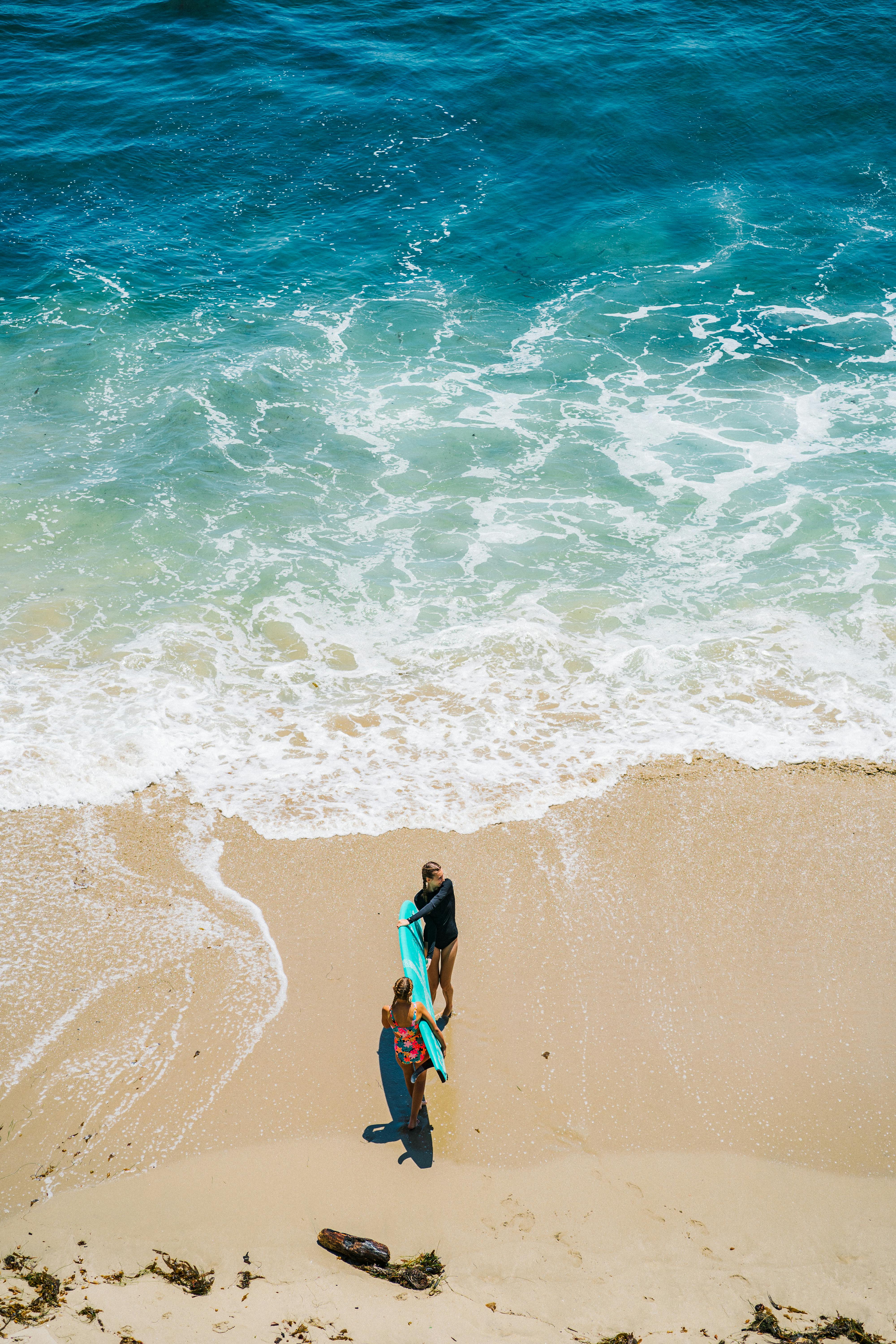 Two surfers carrying surfboards across a sandy shore with vibrant blue ocean waves in the background.