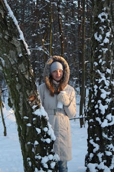 A young Caucasian woman in a winter forest holding a thermos, wrapped in a warm coat, surrounded by snow.