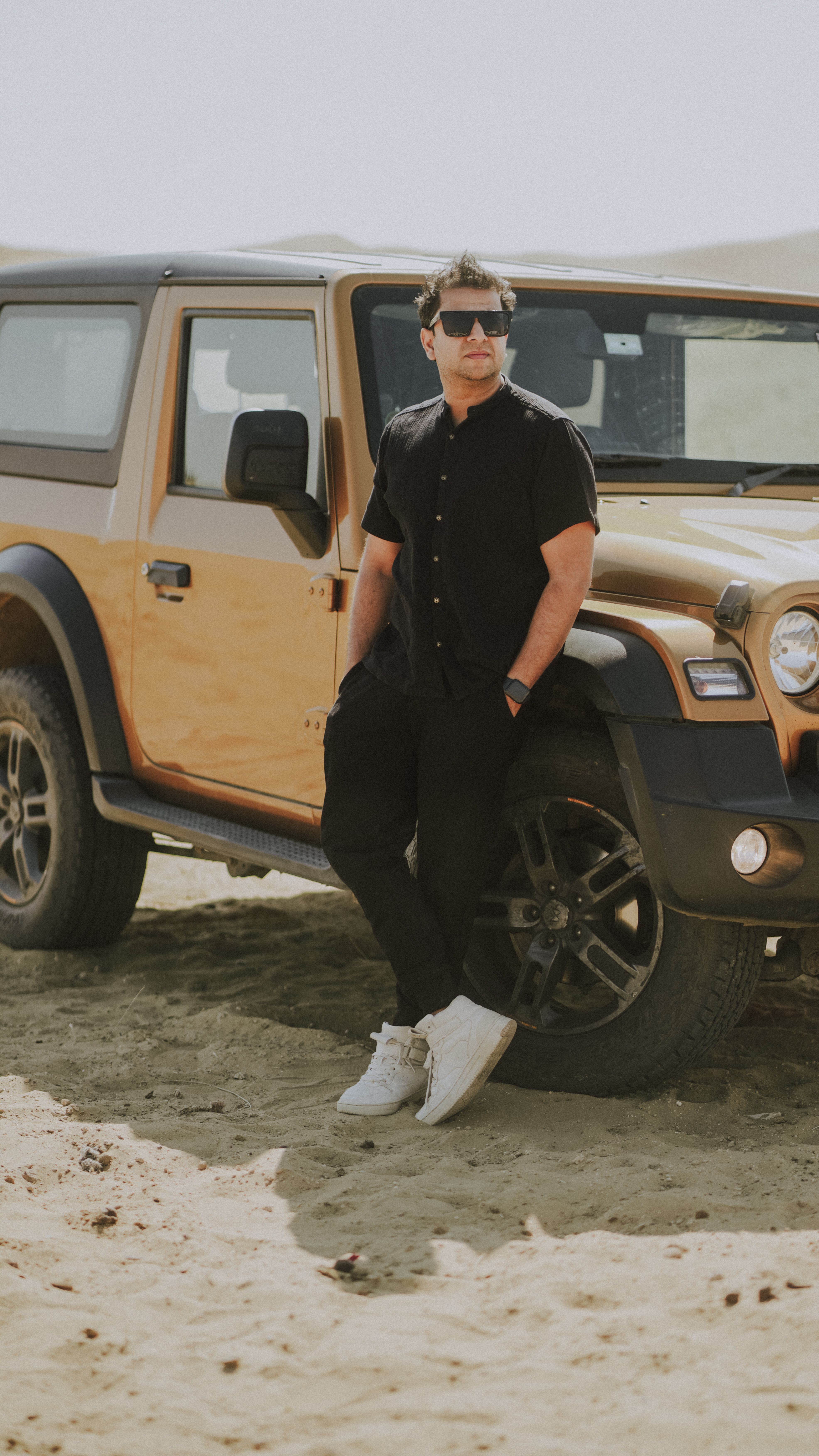 A man in sunglasses leans against a parked SUV in a sunny desert setting, embodying adventure and leisure.