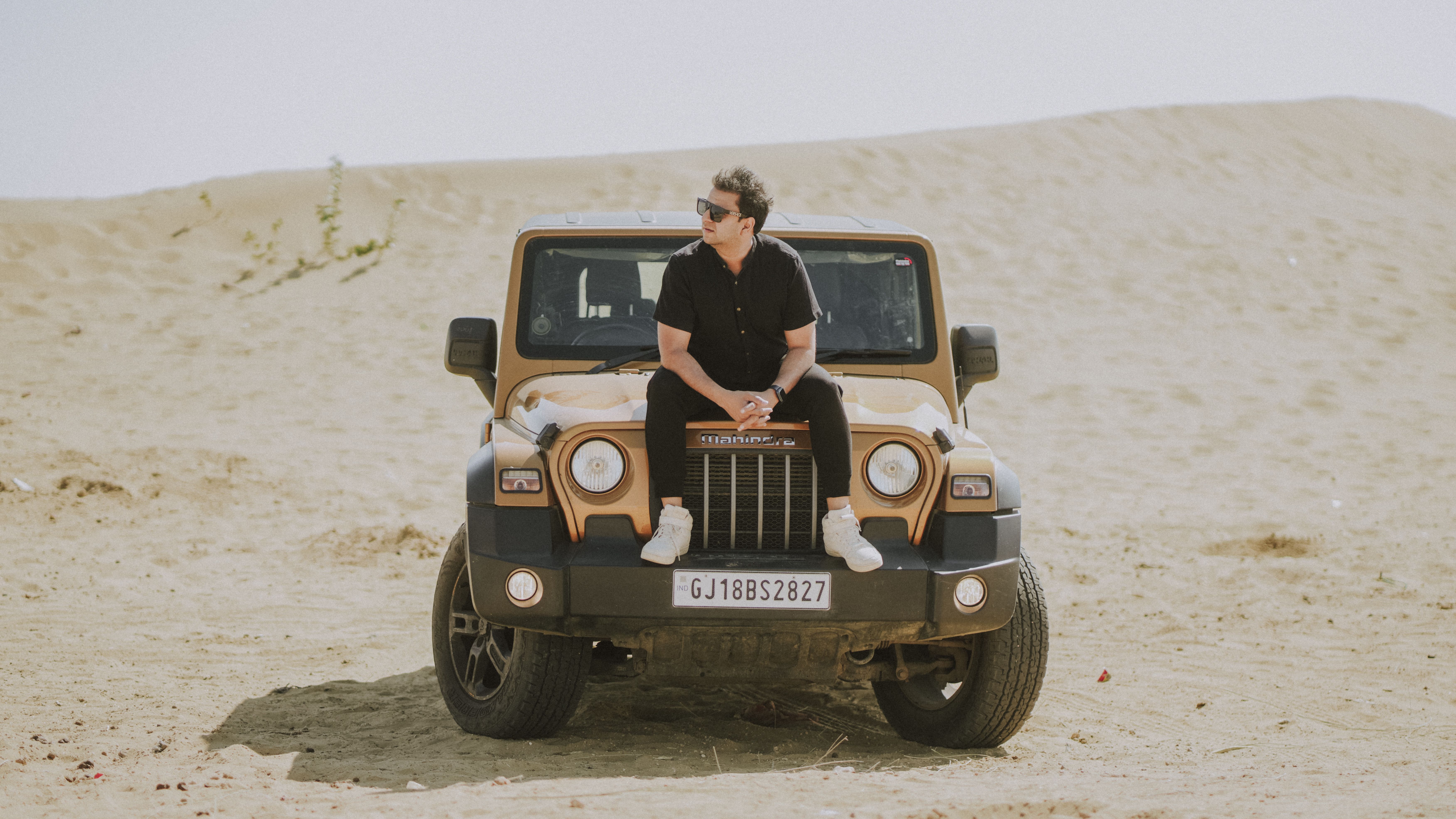 A man sits on a modern SUV parked in a sandy desert, wearing sunglasses and casual attire.
