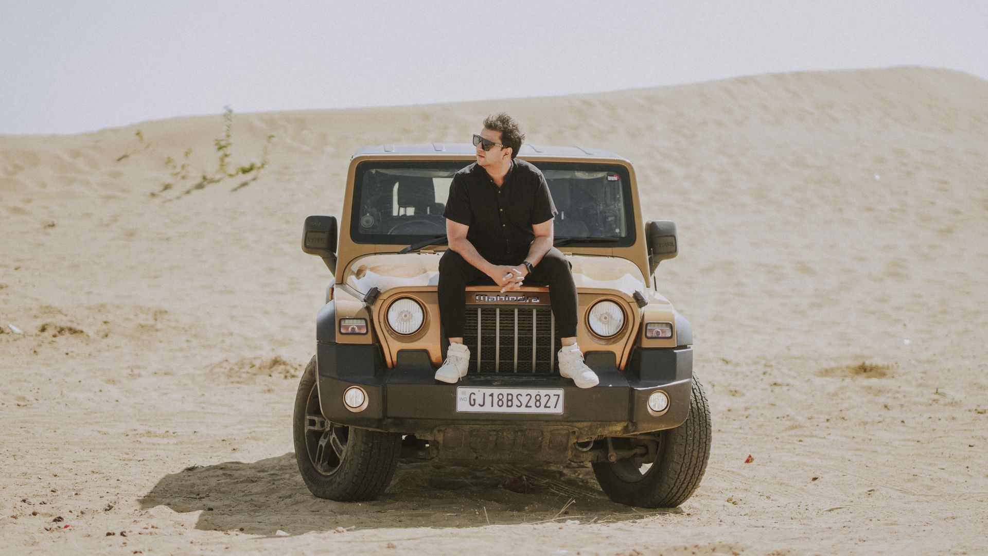 A man sits on a modern SUV parked in a sandy desert, wearing sunglasses and casual attire.
